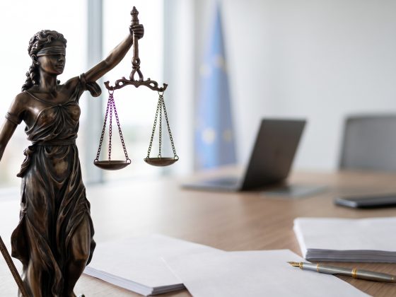 Bronzefigur der Justitia mit Waage und Schwert auf einem Tisch mit Papieren, Stift, Laptop und Stuhl im Hintergrund