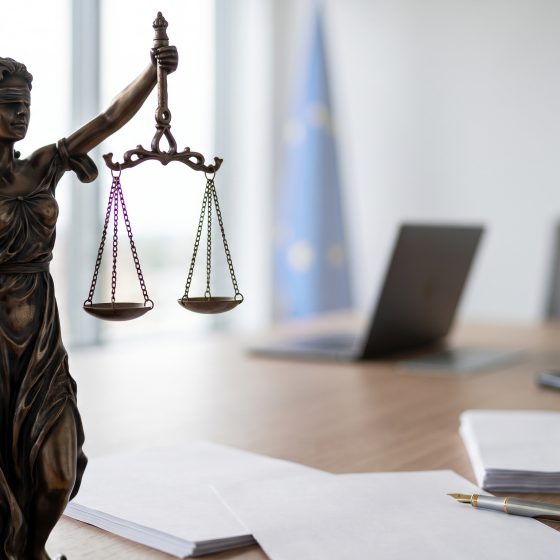 Bronzefigur der Justitia mit Waage und Schwert auf einem Tisch mit Papieren, Stift, Laptop und Stuhl im Hintergrund